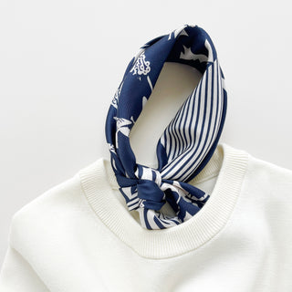 Navy blue and white patterned silk scarf featuring mixed floral, polka dot, and animal print motifs on a white sweater against a light background