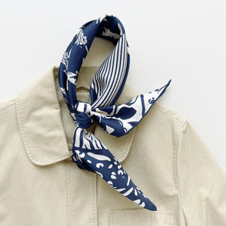 Navy blue and white patterned silk scarf featuring mixed floral, polka dot, and animal print motifs draped over a beige coat on a light background