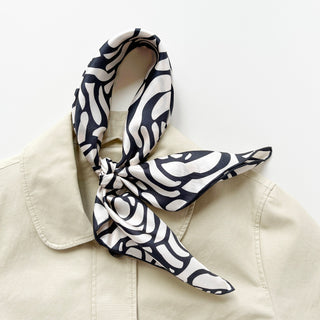 women's silk scarf with charcoal black abstract rose pattern on cream white background, elegantly knotted on a beige coat