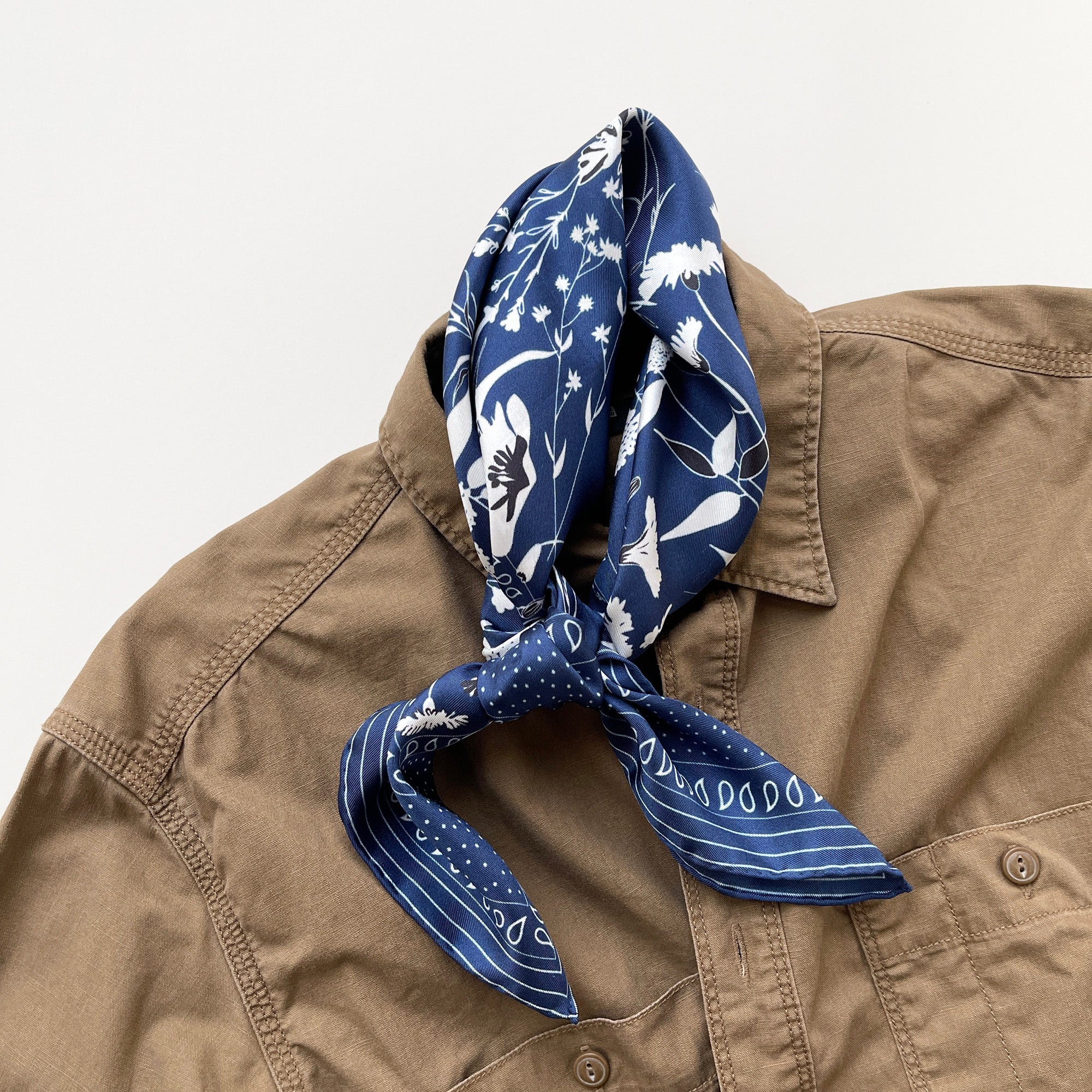 a rich blue men's silk scarf featuring botanic leafy print and hand-rolled hems knotted as a neckerchief, paired with a khaki shirt
