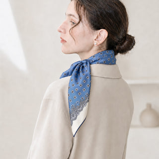 Woman wearing a cornflower blue vintage medallion 65cm silk square scarf tied at the back of her neck, styled with a low bun and ivory jacket.