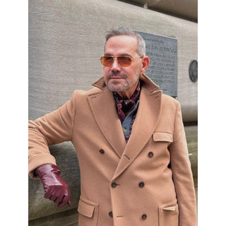 Man wearing a burgundy paisley silk square scarf by LuluLane in a camel coat and sunglasses standing against a textured wall.