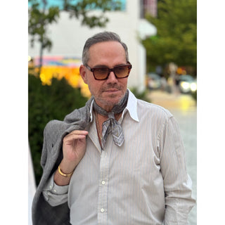 Man wearing a grey and taupe tone patterned silk square from LuluLane in a striped shirt and sunglasses outdoors with blurred background showcasing a polished smart casual look.