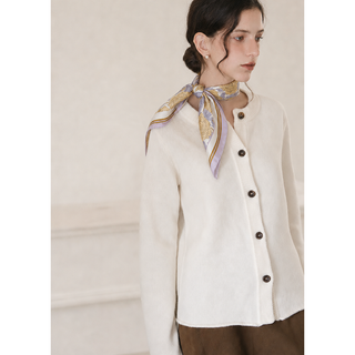 Woman wearing a lilac peony print silk neck scarf tied elegantly at the side of the neck, styled with a cream-coloured button-up cardigan and pearl earrings. 