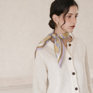 Woman wearing a lilac peony print silk neck scarf tied elegantly at the side of the neck, styled with a cream-coloured button-up cardigan and pearl earrings. 