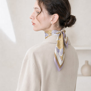 Woman wearing a lilac Peony Bloom silk neck scarf tied neatly at the back of the neck, styled with a cream coat and pearl earrings.