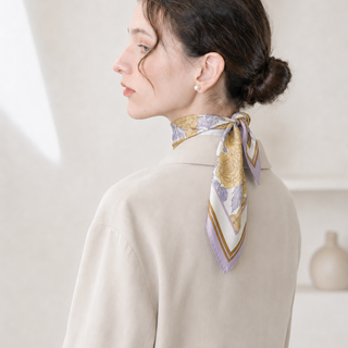 Woman wearing a lilac Peony Bloom silk neck scarf tied neatly at the back of the neck, styled with a cream coat and pearl earrings.