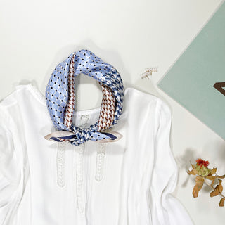 Blue silk bandana with geometric patterns tied at the neck of a white blouse, displaying versatile styling of the unisex scarf