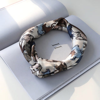 Luxurious silk bandana scarf with elegant horse motif in cream, blue, and brown tones, artfully folded in circular shape on open book pages