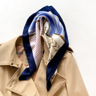 Silk bandana scarf with geometric pattern in blue, pink, and cream with navy border, styled on beige jacket collar for elegant accessory look.