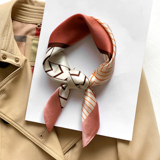 Silk scarf in pink and coral tones with geometric patterns, elegantly knotted and displayed on white paper over a beige trench coat