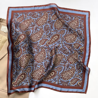 Silk scarf with blue background featuring intricate brown paisley pattern and bordered with brown trim, elegantly displayed on cream surface