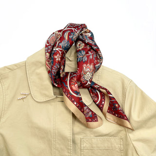 Red vintage silk scarf with ornate floral pattern and golden border draped elegantly around the collar of a beige jacket