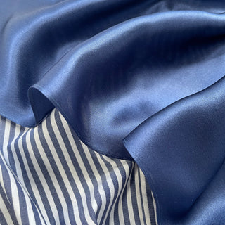 Navy blue silk scarf with smooth, lustrous finish draped over striped blue and white fabric, showing the scarf's soft texture and rich color
