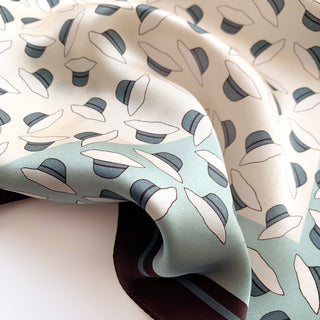 Luxurious silk scarf with playful hat pattern in cream and soft blue, featuring navy blue hat details with a smooth, shiny texture.