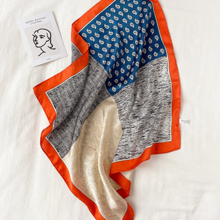 Square silk neckerchief with blue paisley pattern, cream paisley design, black and white textured print, and vibrant orange border, displayed on white surface