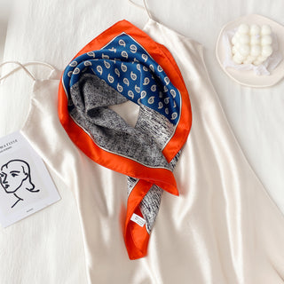 Square silk neckerchief with orange border, blue paisley pattern, and black and white patchwork design, displayed on white fabric