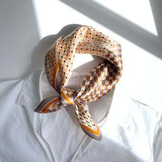 Silk scarf with geometric patterns in peach, orange, and white tones, elegantly tied around a white collar, showcasing luxurious texture and versatility.