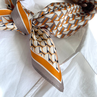 Silk bandana with geometric pattern in orange, gray and cream tones, elegantly knotted on white fabric showing its lustrous texture