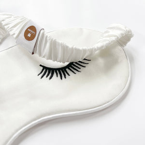 a white silk eye mask featuring black eyelash embroidery and elastic strap