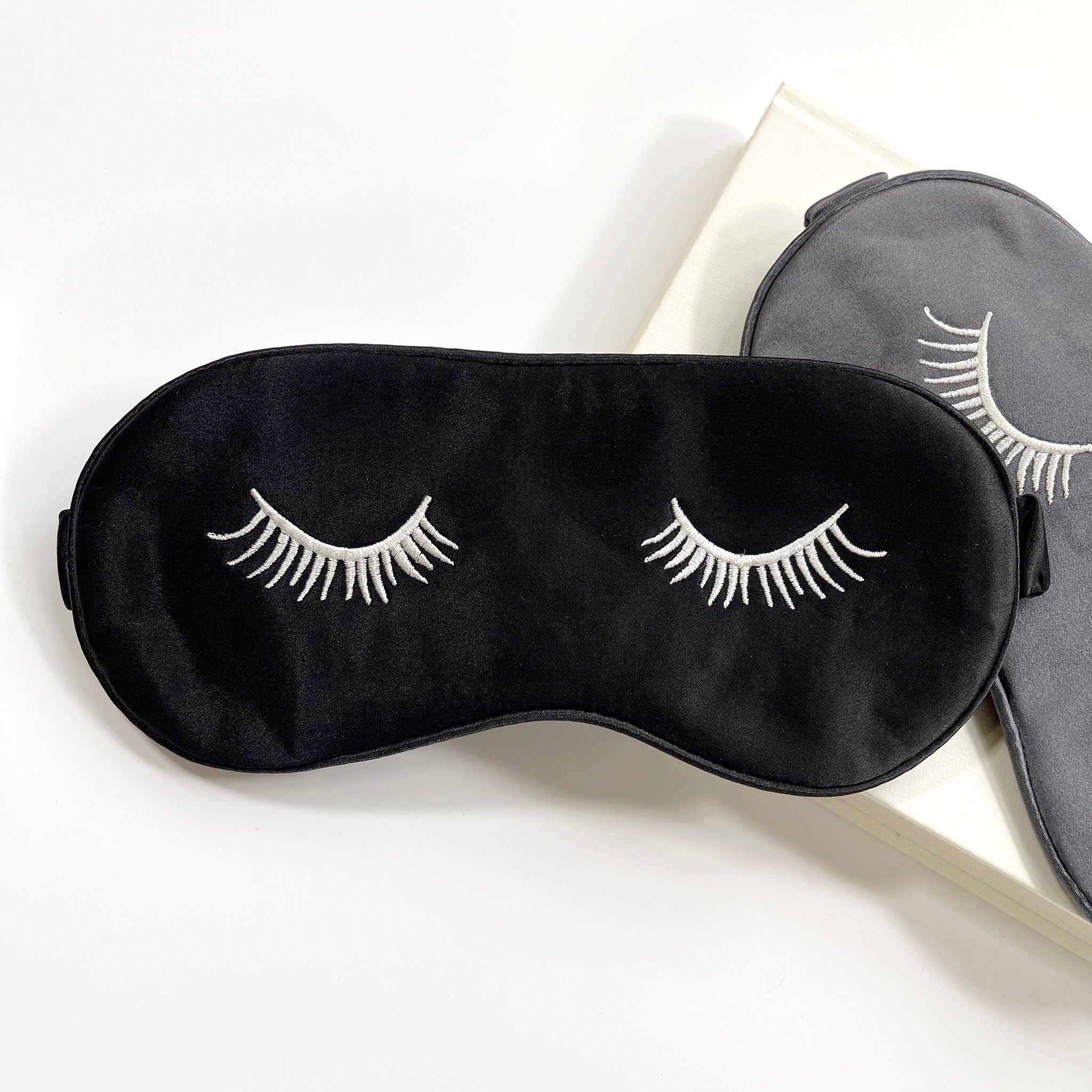 two silk eye masks featuring eyelash embroidery in black and grey colors