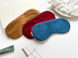 Luxury silk sleep masks in three colors - caramel gold, burgundy red, and teal blue - with elegant gold trim, displayed on white surface.
