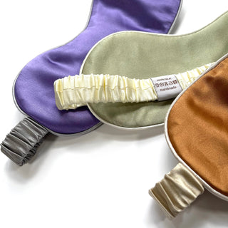 Luxury silk sleep masks in purple, sage green, and caramel gold with elastic bands and 100% silk label, displaying double-sided design on white background