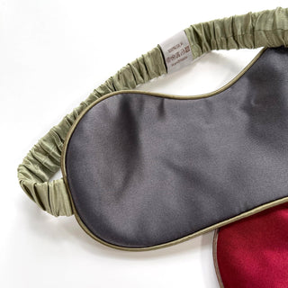Double-sided silk sleep mask with gray and red sides, elastic sage green band, and 100% silk label, showing luxurious texture and craftsmanship.