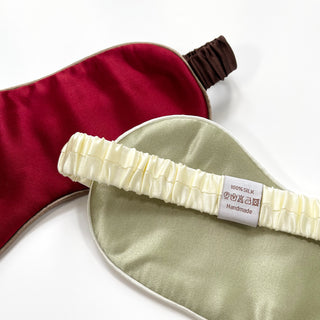 Two luxury silk sleep masks with elastic bands - one deep red with brown strap and one sage green with cream strap, showing 100% silk label