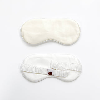Luxury cream-colored silk sleep mask shown from front and back views, featuring soft fabric texture and adjustable elastic band with brand tag.