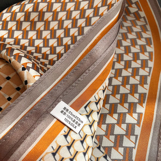 Silk scarf with geometric pattern in orange, gray, and cream tones, showing intricate triangular designs and border details with product tag